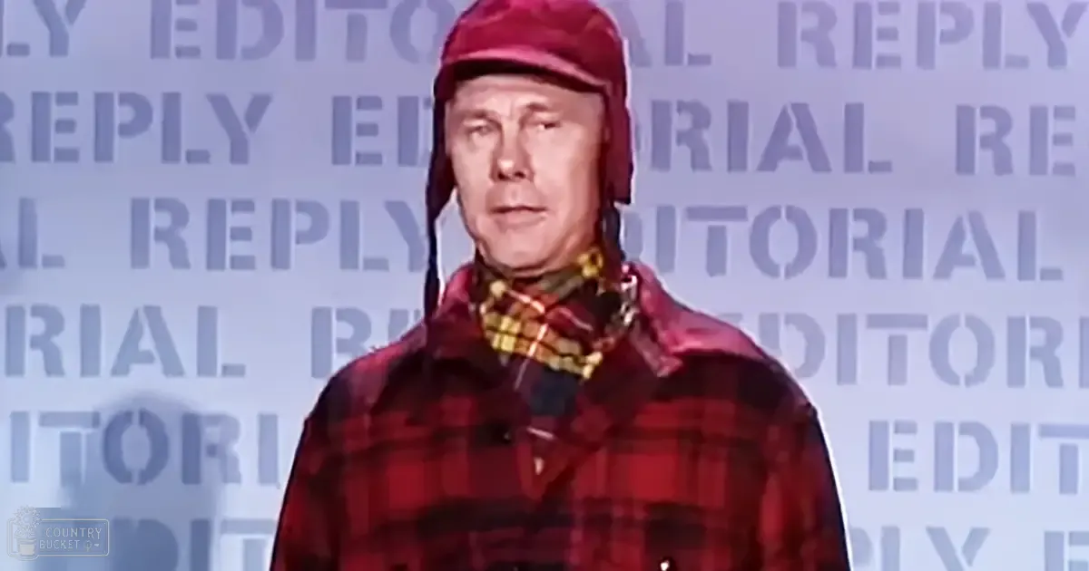 A middle-aged adult male in a mid-20th-century style red plaid jacket with dark accents and shiny red helmet, accessorized with a yellow and black plaid scarf. The backdrop has repeated text reading 'EDITORIAL REPLY' on an indoor setting.