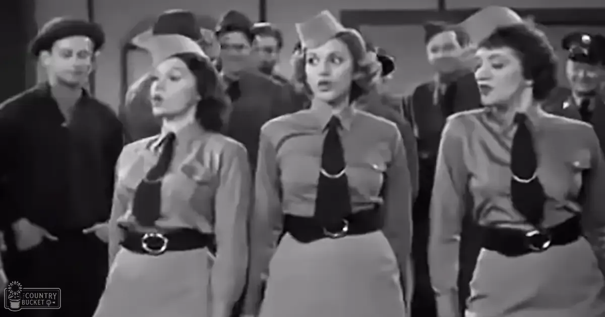 A group of women in World War II-era uniforms wearing long-sleeved shirts with collars and ties, standing indoors alongside two adult males in dark shirts and pants. The uniforms suggest a historical or reenactment setting.