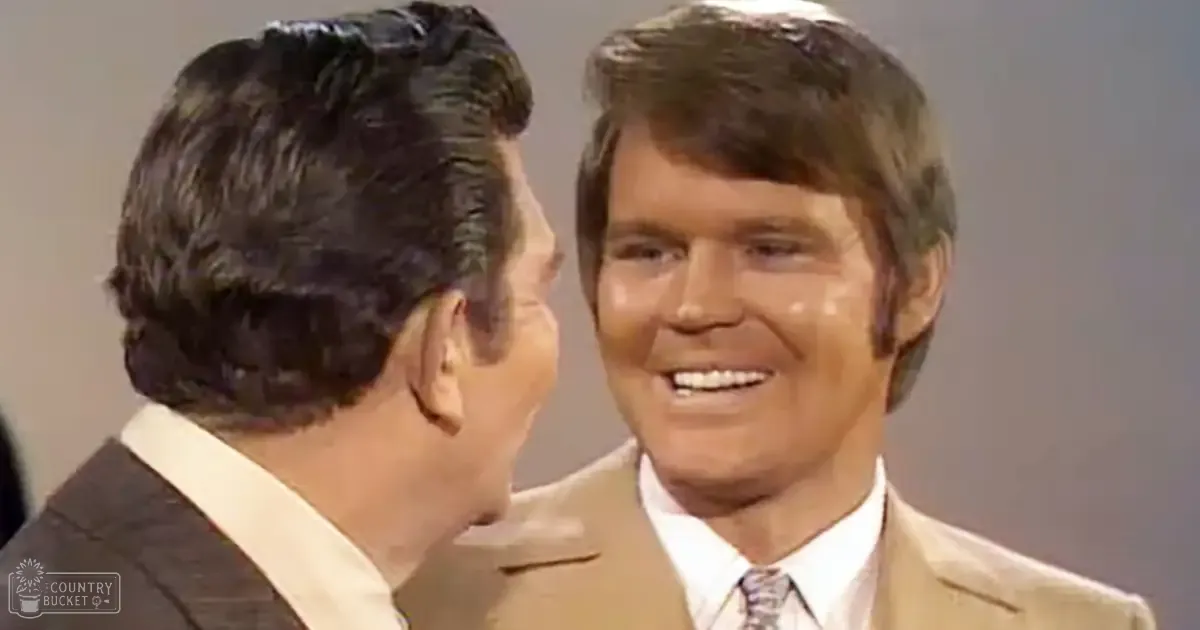 An adult male singer wearing a dark suit jacket over a white shirt stands next to another middle-aged man in a light-colored suit and patterned tie, both set against an indoor backdrop with even lighting.