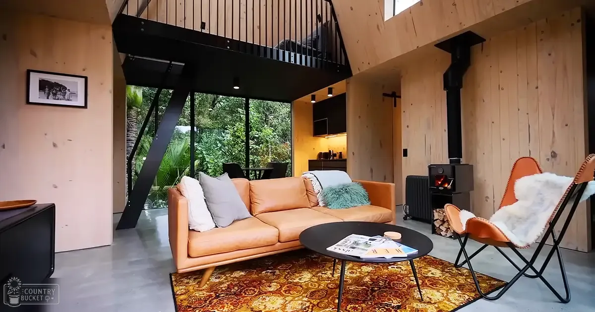 A cozy modern living room with light brown leather sofa, orange armchair, wooden wall paneling, black coffee table holding magazines, and a view of greenery outside.