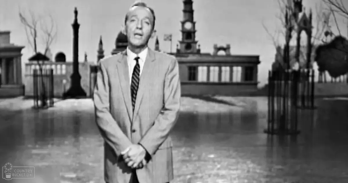 A middle-aged adult male wearing a light gray wool suit with a striped tie, standing outdoors in what appears to be a wet town square or park setting.