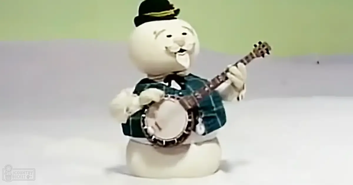 A snowman figurine wearing a green hat and playing a banjo with a white drumhead, set against a plain indoor background.