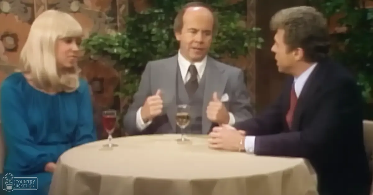 A talk show set with three adults dressed formally, seated around a round table with glasses of liquid and decorative greenery in an indoor studio setting.
