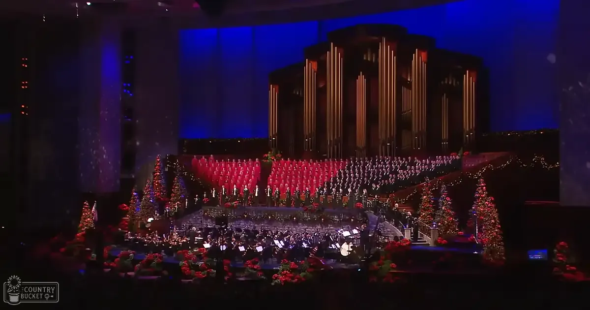 A Christmas concert with a large organ, choir in red robes, and orchestra in dark uniforms indoors under artificial stage lights.