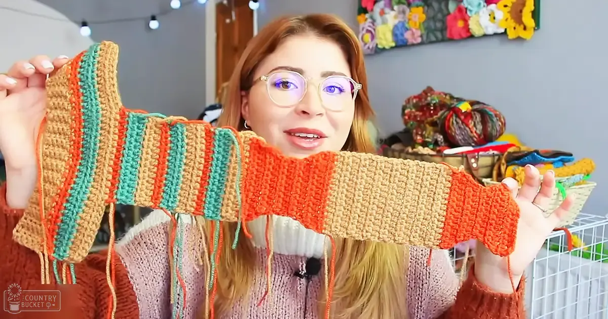 A young adult female wearing a pinkish-beige cable-knit sweater with long sleeves and a high neckline, accessorized with glasses, in an indoor crafting or hobby area with string lights and colorful wall hangings.