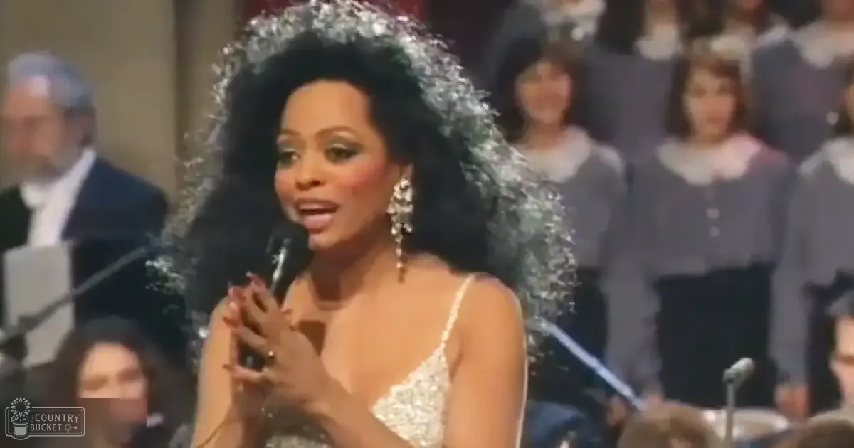 An adult female performer in a formal indoor concert setting, wearing an intricate white sleeveless dress with beadwork and sequins, accompanied by large dangling earrings; other adults are partially visible in the background, suggesting a choir or ensemble setup.