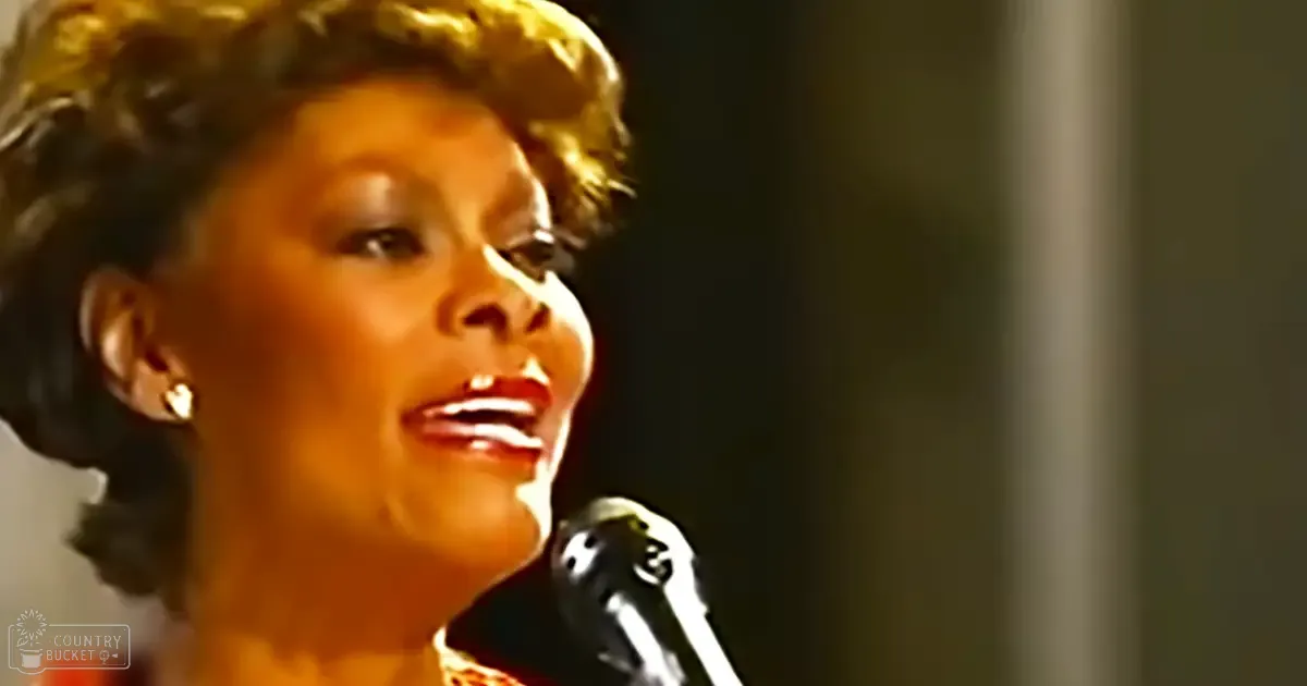 An adult female singer in an indoor setting, wearing a red and white patterned garment with 'Madly Odd' printed on it, holding a black microphone.