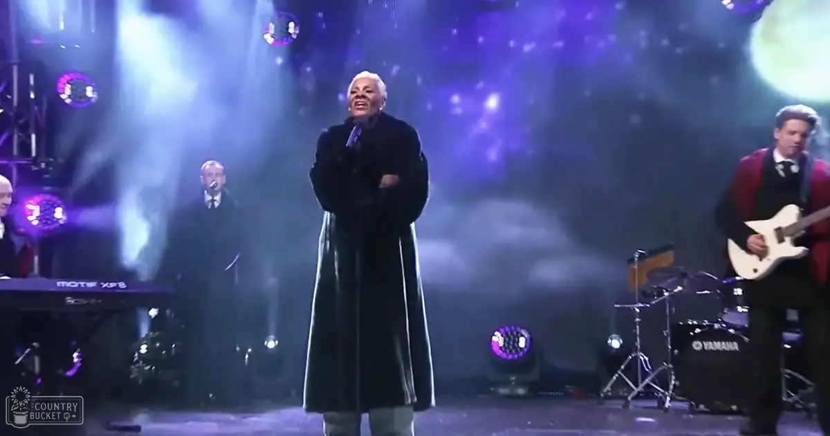 An older adult male performer in a black coat with white hair, surrounded by background musicians playing instruments under stage lights creating a dramatic atmosphere.