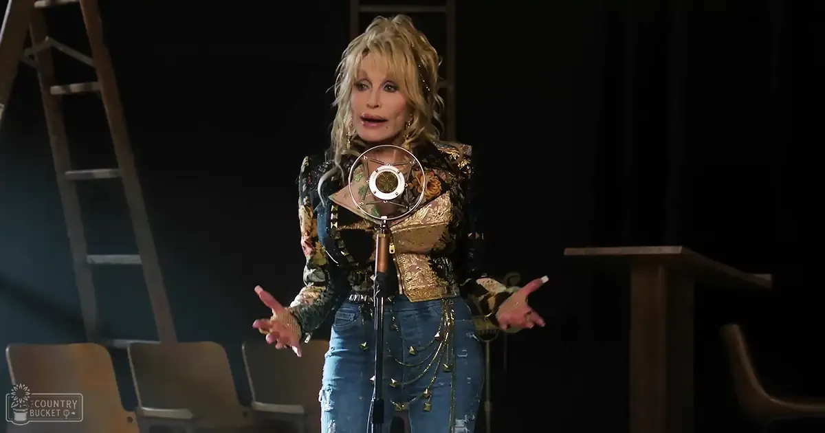 Middle-aged female performer wearing a patterned floral and paisley jacket with intricate embroidery, gold chains, and blue jeans with distressed details and star-shaped decorations on stage under focused lighting.