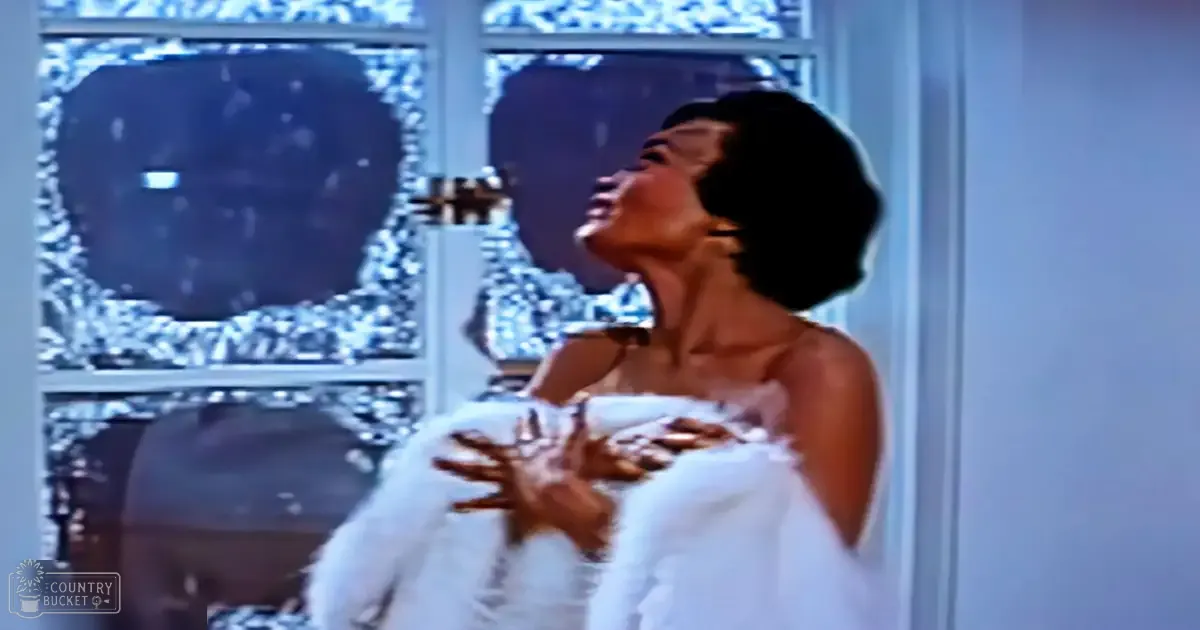 A woman singing indoors, wearing a white, feather-like textured garment with an elegant mid-20th-century style. The setting is decorated for winter holidays with Christmas trees and snow-covered trees visible through the window.