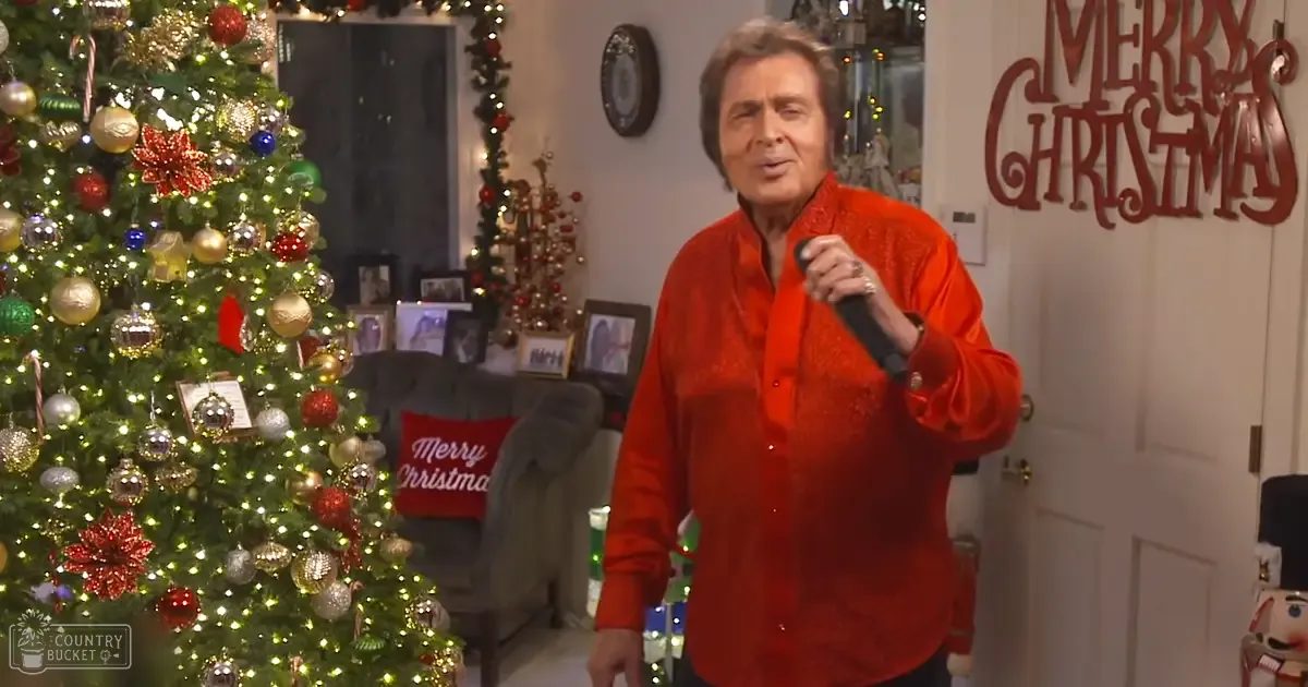 A festive indoor scene with an older adult male wearing a bright red, collared long-sleeved shirt in front of a decorated Christmas tree and armchair.