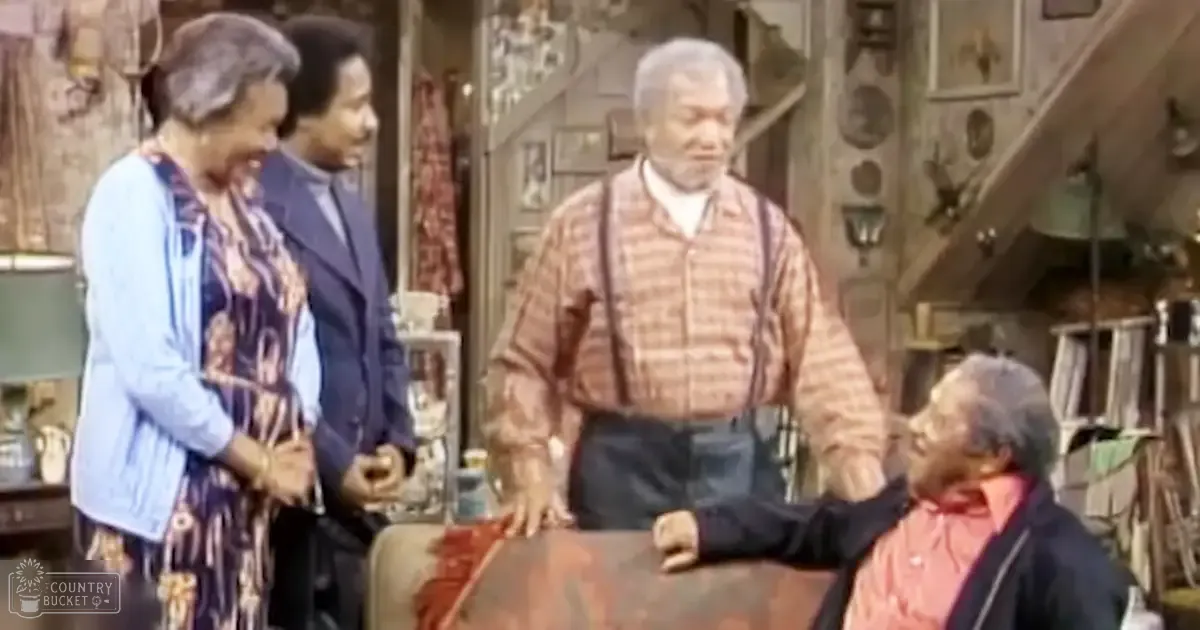 An older adult male standing with two other adults, one female to the left wearing a patterned dress with a light blue cardigan, another male in the center in a striped shirt and suspenders, and an older female on the right in a pink jacket with a dark scarf draped over her shoulders. They are in what appears to be a home workshop or garage setting with various tools and shelves visible.