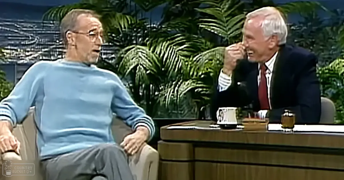 A talk show set with two middle-aged male hosts, one wearing a light blue sweater and the other in a dark suit jacket over a white shirt with red tie, seated at a wooden desk adorned with green foliage in an indoor setting designed to mimic a tropical environment.