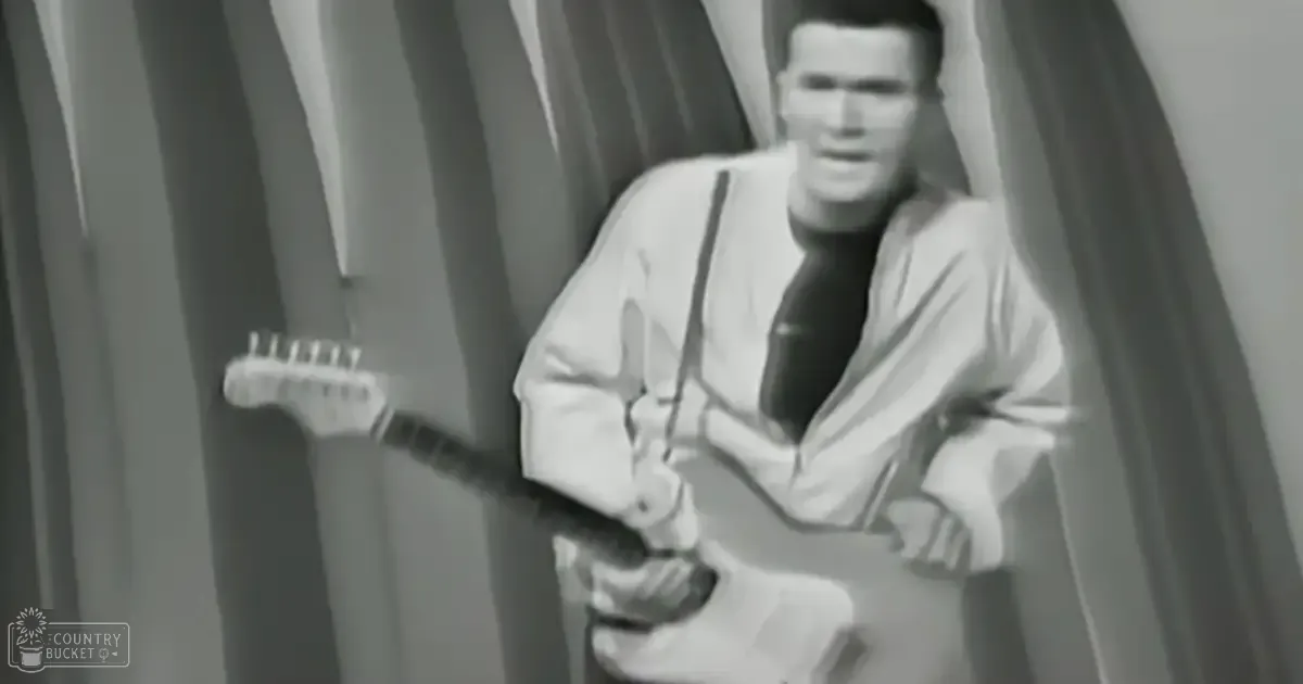 A young adult male plays an electric guitar in a studio setting, wearing a white shirt and black tie.
