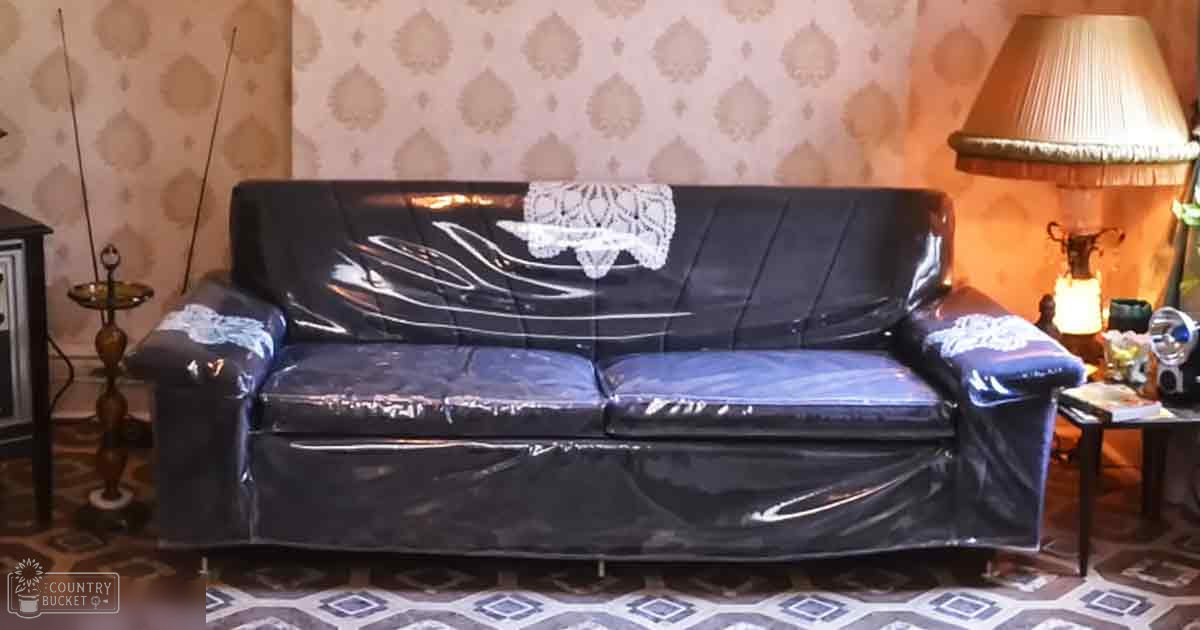 An ornate wallpapered living room scene with a black leather sofa covered in plastic wrap and small side tables holding decorative items, illuminated by artificial lighting from a floor lamp and candle holder. The setting suggests evening or night due to the use of lights.