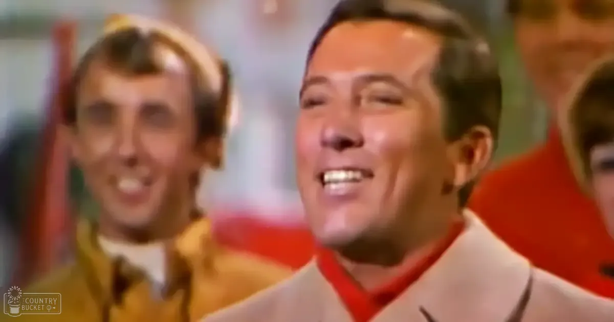 Middle-aged man in a light-colored suit jacket, seated indoors with other individuals wearing casual attire; festive or holiday setting