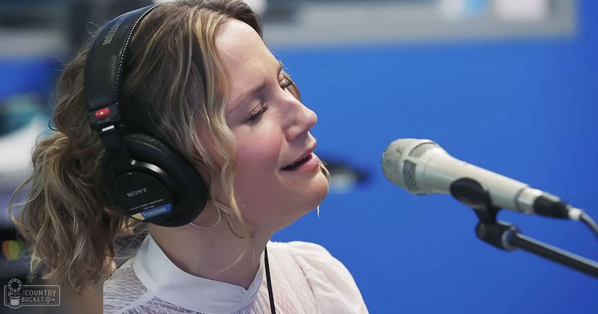 A young adult female in a recording studio wearing a white top with a mesh-like pattern, long sleeves, black Sony headphones, and standing against a blue backdrop.