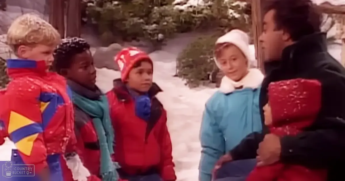 Children in a snowy outdoor setting with adults; children wearing red jackets, blue accents, and knit hats, adult male in a black jacket with red accents on shoulders and back;