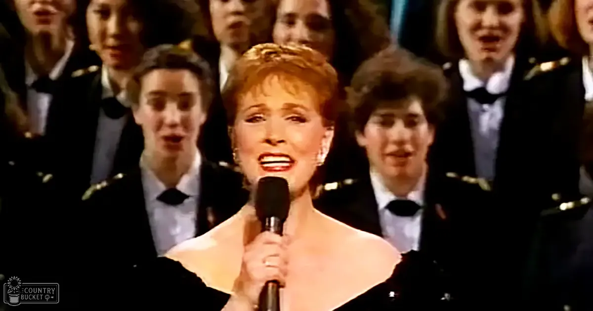 A middle-aged female performer singing in front of a choir dressed in military-style attire under stage lighting, suggesting an evening performance.