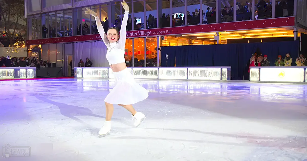 A young adult female figure skating at an ice rink in Bryant Park's Winter Village during a festive evening with holiday lights.