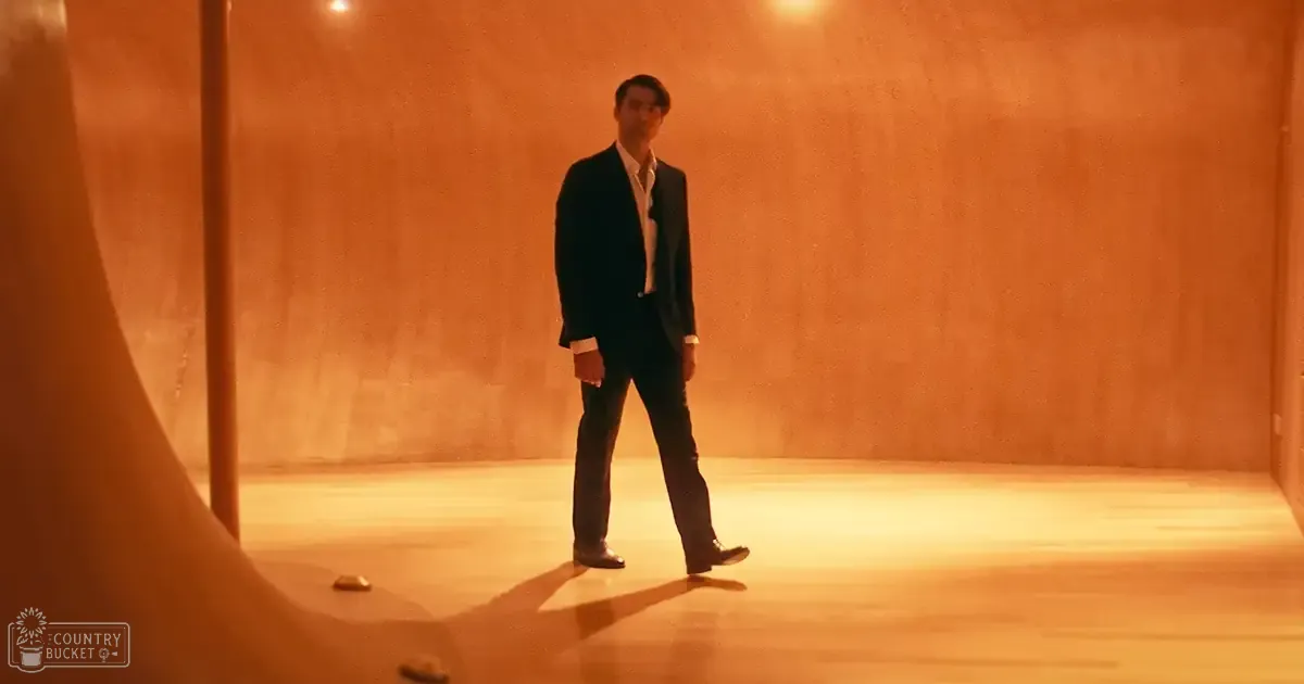An adult male singer wearing a dark suit jacket and trousers with a white dress shirt, standing indoors under dramatic orange-toned lighting.