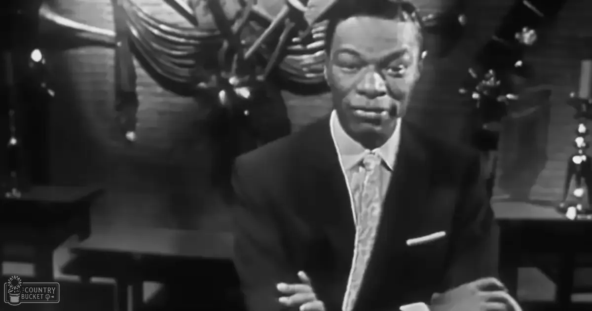 A middle-aged man in a dark suit with a white pocket square and patterned tie stands indoors, possibly at a social gathering or performance.