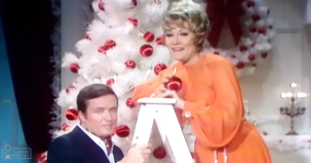 A middle-aged woman wearing an orange dress interacts with a middle-aged man in a dark suit jacket, both dressed formally for what appears to be a festive event indoors, with Christmas decorations and props visible around them.
