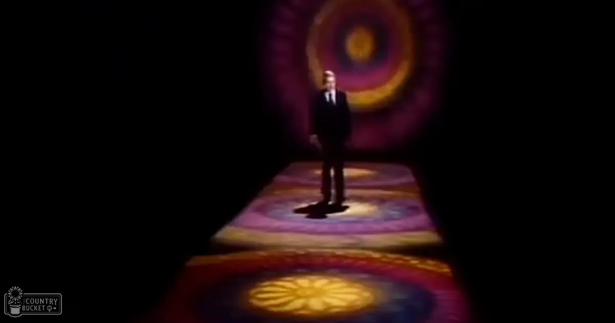 An adult male wearing a classic dark suit and tie stands before a colorful, patterned backdrop in what appears to be an indoor performance venue with stage lighting.
