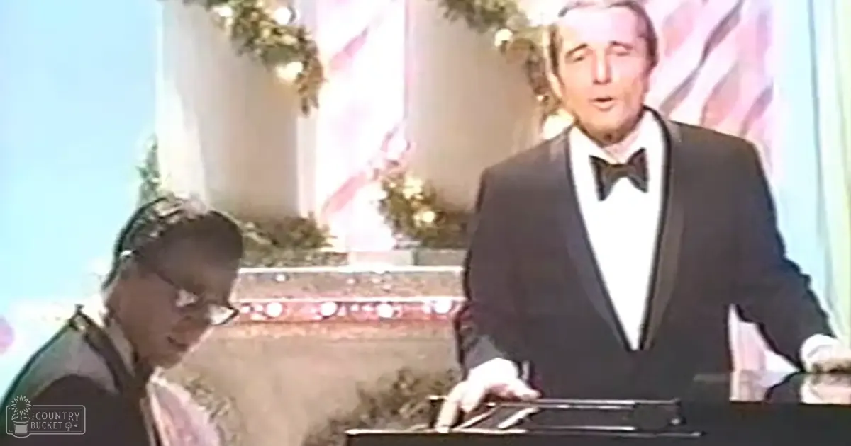 A middle-aged man in a formal black tuxedo and bow tie stands at an awards ceremony indoors with decorative elements around him.