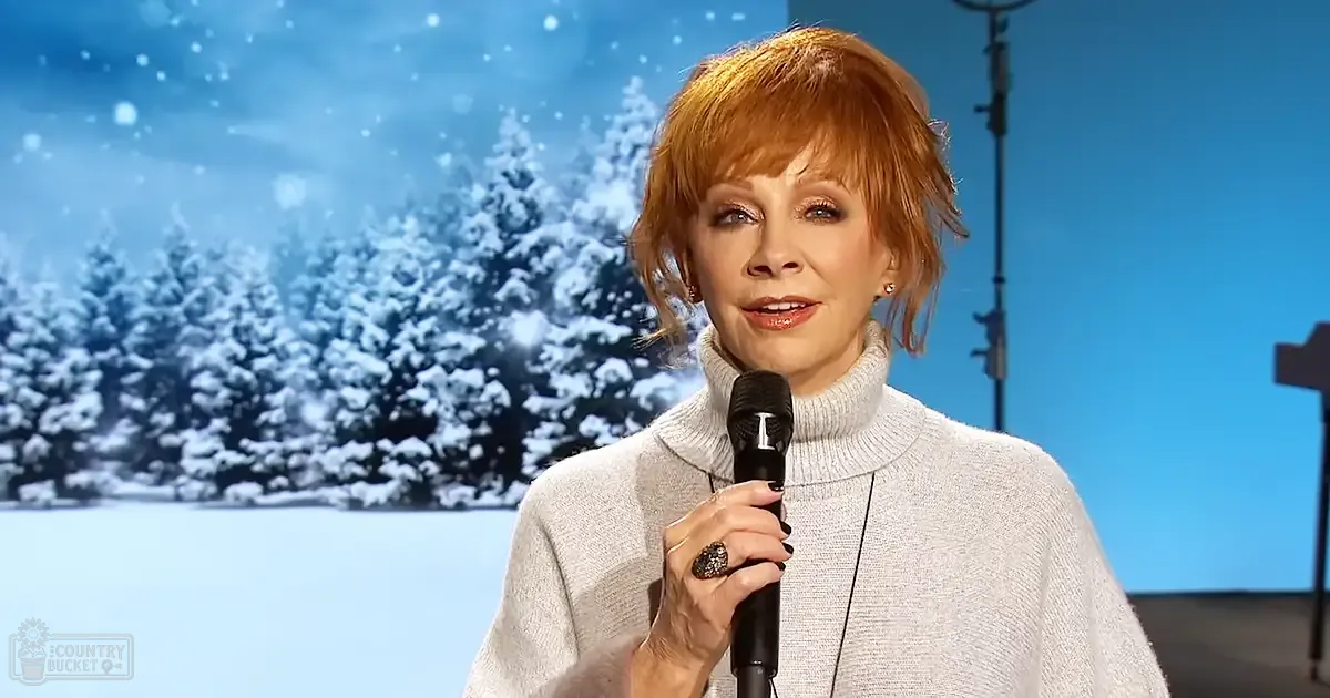 Middle-aged adult female wearing a light gray turtleneck sweater in an indoor winter setting with snow-covered trees and blue sky background.