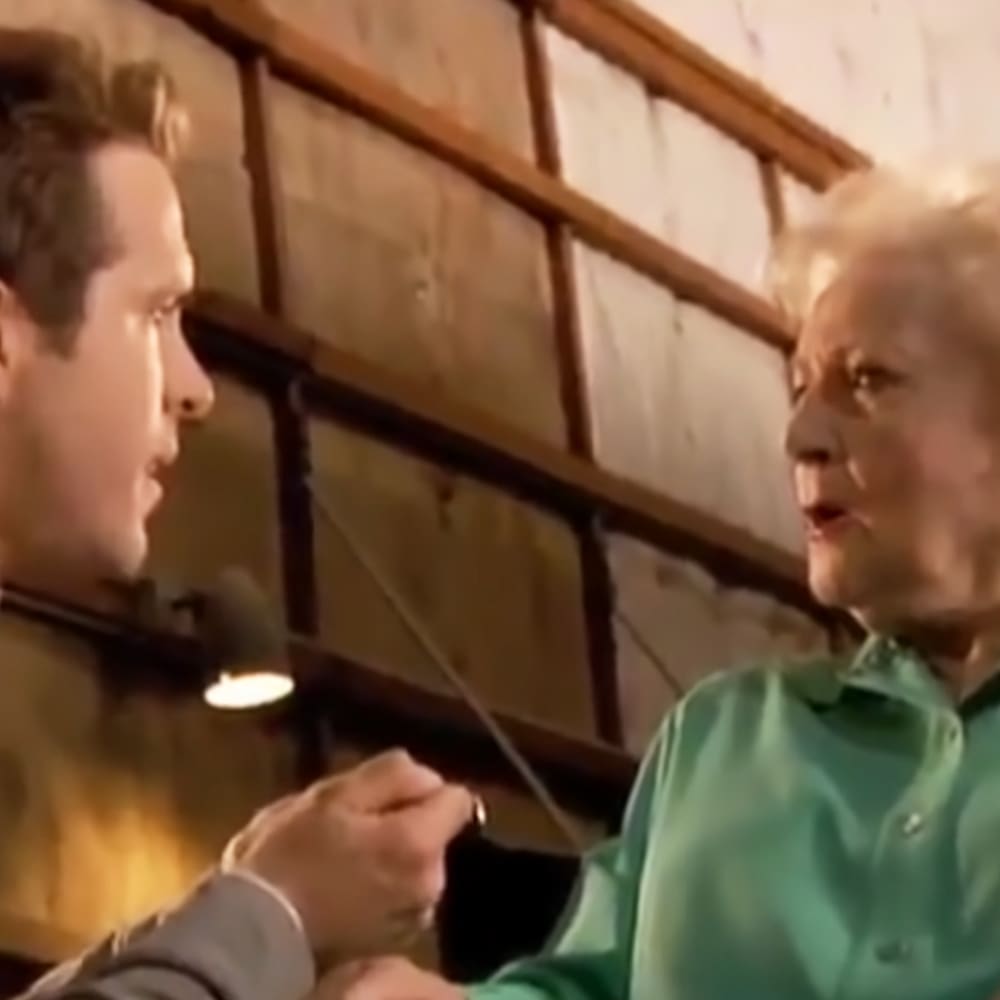Two middle-aged to older adults conversing indoors, one wearing a light-colored long-sleeved shirt and the other in a teal button-up shirt with natural lighting suggesting late afternoon or early evening.