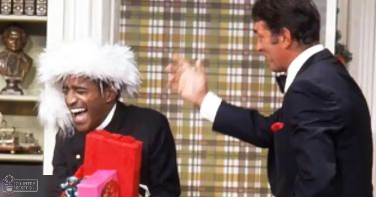 A comedic scene from a television show or film set indoors during winter, with two men in black suits with white lapels against a plaid wallpaper backdrop.