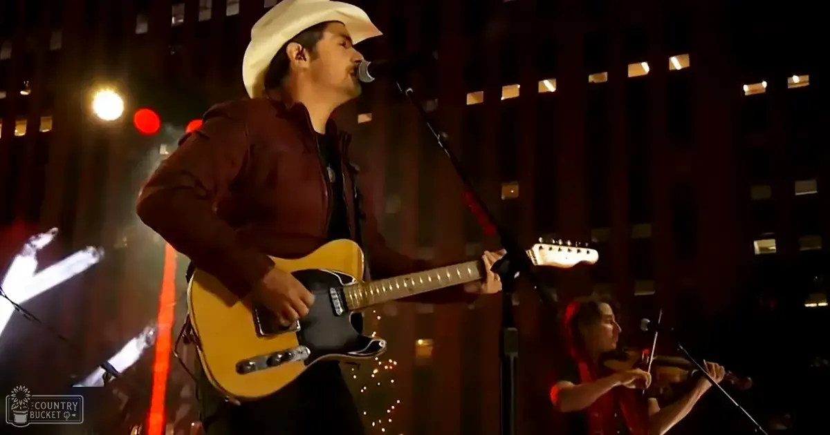 An adult male performer wearing a dark brown jacket with a high collar and white cowboy hat stands on an urban stage at night, accompanied by another young adult female musician playing the violin.