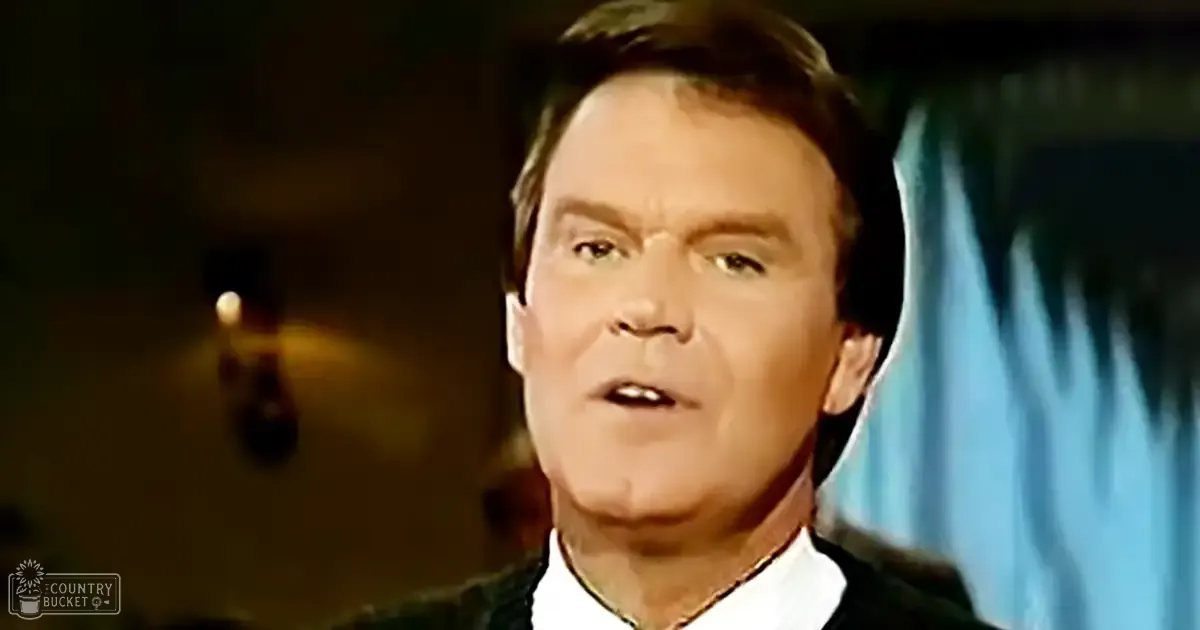 A middle-aged adult male in a relaxed, dark-colored sweater with a white collared shirt underneath, indoors with dim lighting and a blue curtain visible.