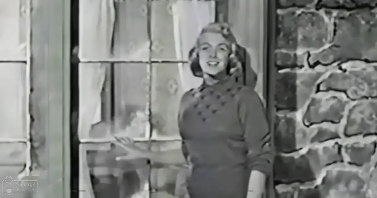 An adult female wearing a dark, heart-patterned long-sleeved dress with a fitted bodice and flared skirt stands indoors near stone walls under bright daylight.