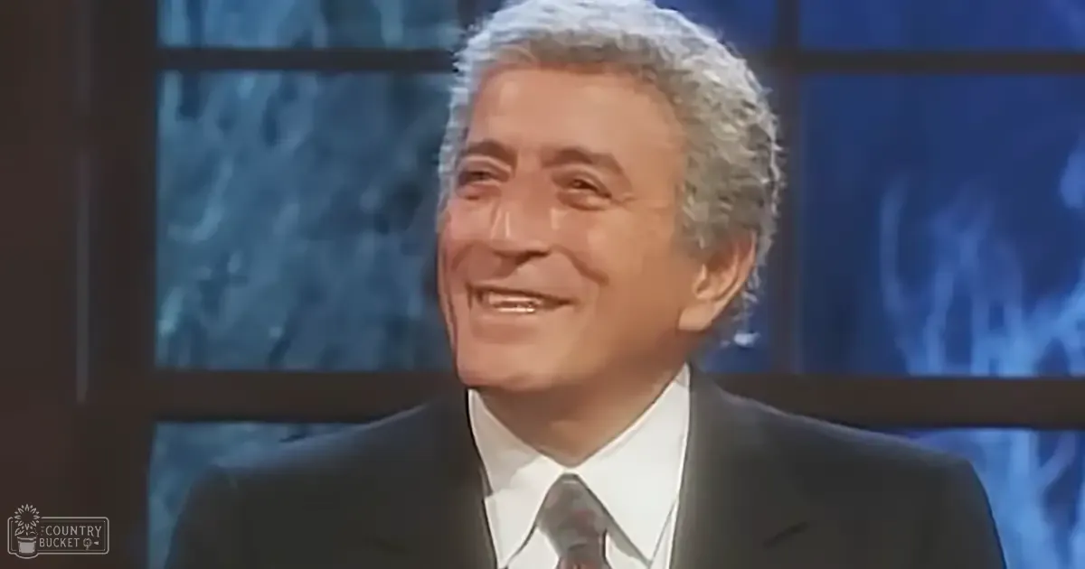 An older adult male in a studio setting, wearing a dark suit jacket, white dress shirt, and patterned tie, standing against a backdrop of blurred audience with blue lighting.