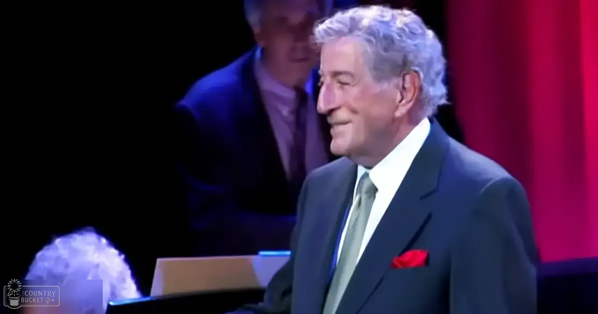 An older adult male performer wearing a dark suit with white shirt, light green tie, and red pocket square, set against a stage backdrop with red curtains.