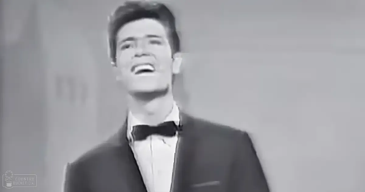 A young adult male wearing a formal suit with a bow tie, standing indoors under artificial light.