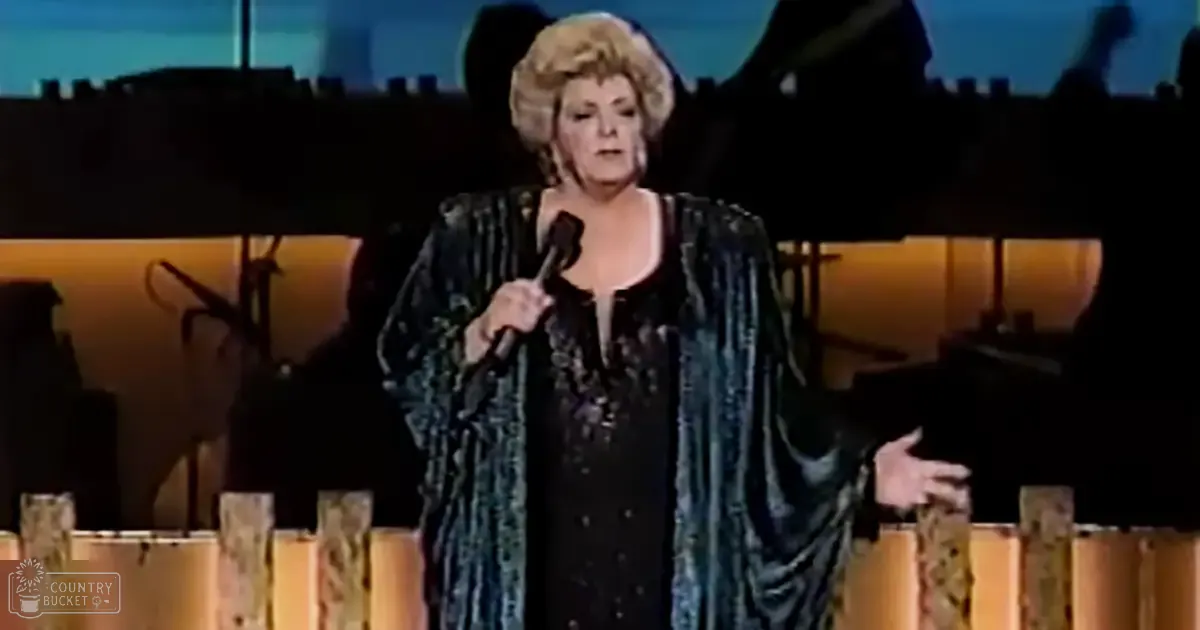 A middle-aged female performer wearing a dark, intricate patterned dress in an indoor stage setting with professional lighting.