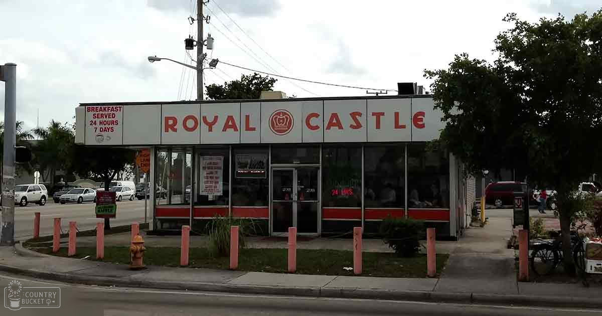 Royal Castle restaurant with large glass windows, pink bollards, trees, and parked cars on a sunny urban street.