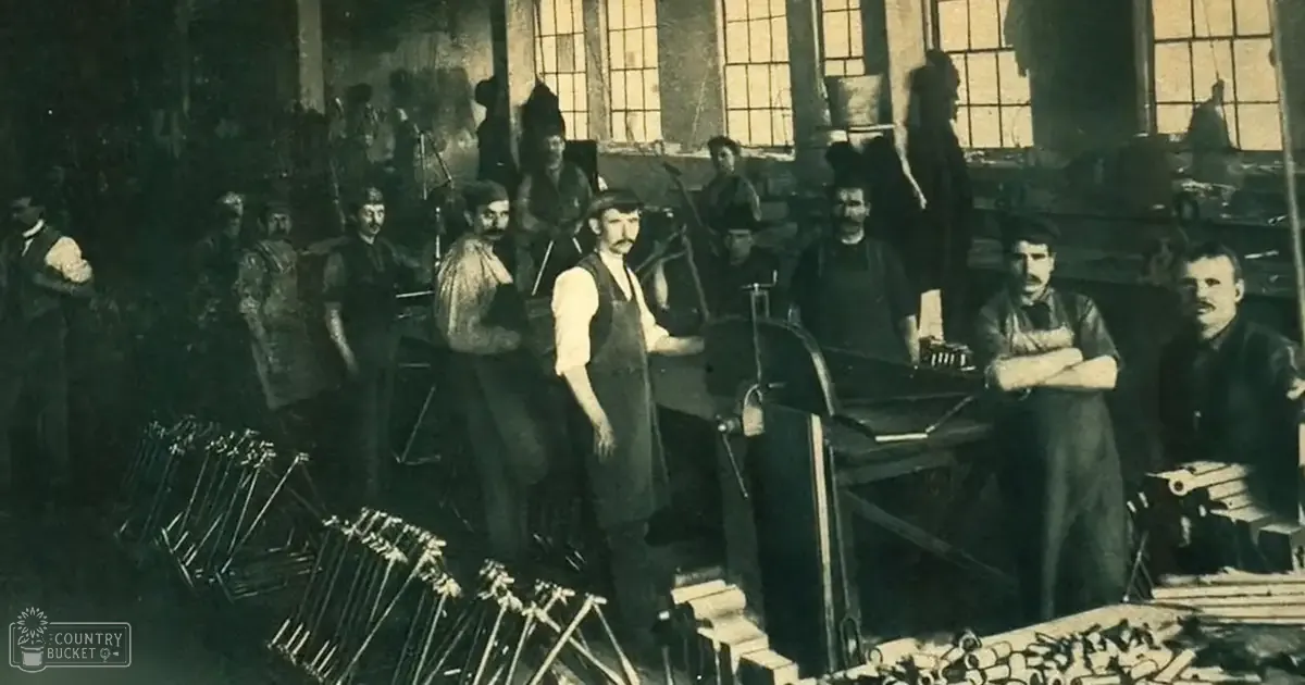 Group of adult males in dark jackets working in a dimly lit industrial space with natural light streaming through large windows and various tools and materials surrounding them, suggesting an older, utilitarian workspace.