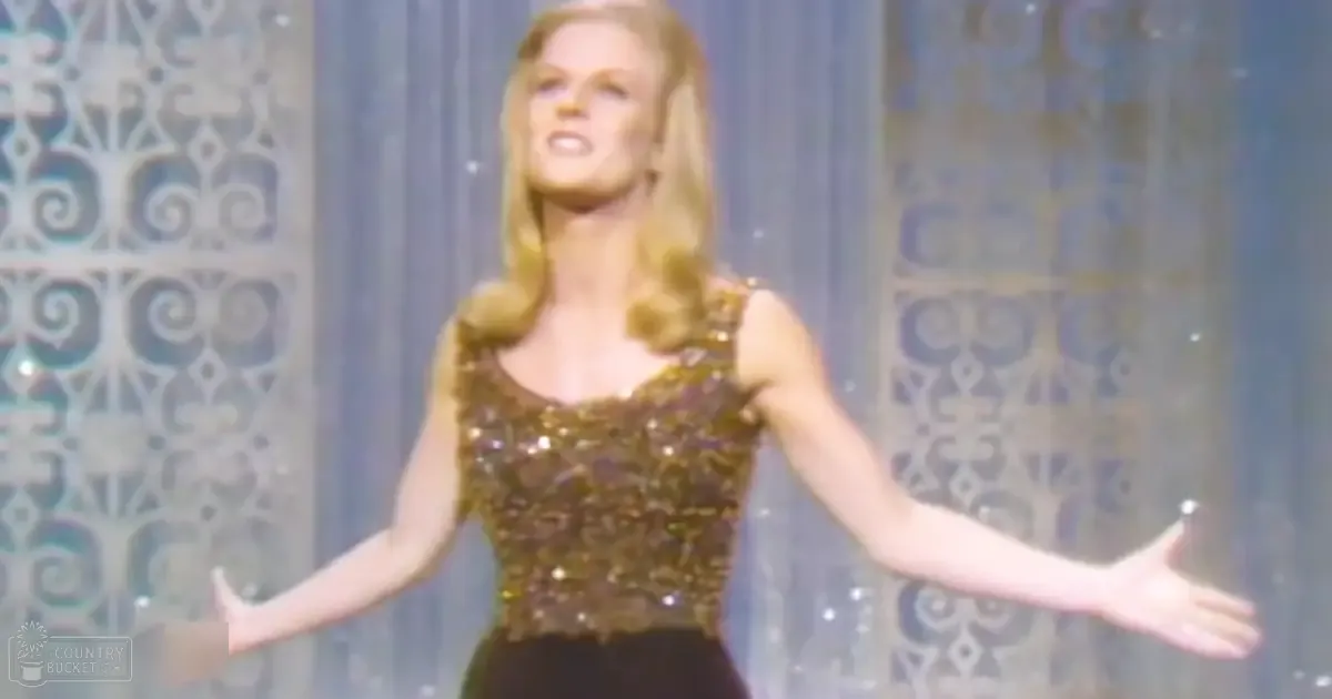 Adult female performer in a sleeveless sequined top and black pants, set against an indoor stage backdrop with intricate patterns. Gold top, black pants, blue-grey background.