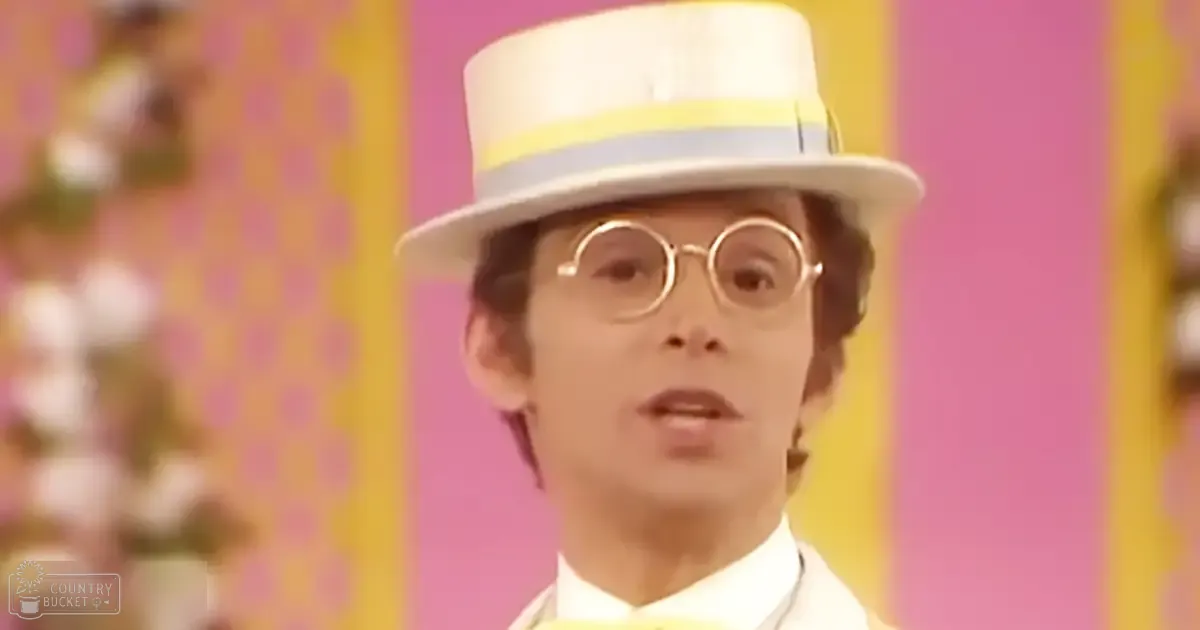 A young adult male in a vintage-style outfit, wearing a white top hat with light blue and yellow stripes, round gold-rimmed glasses, and a high-collared shirt or jacket. The setting is an indoor room decorated with vibrant pink and yellow walls, suggesting a festive atmosphere.