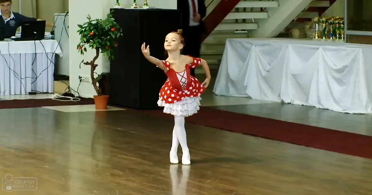 A young female dancer in a red and white polka-dot leotard performs indoors, with bright lighting suggesting daytime.