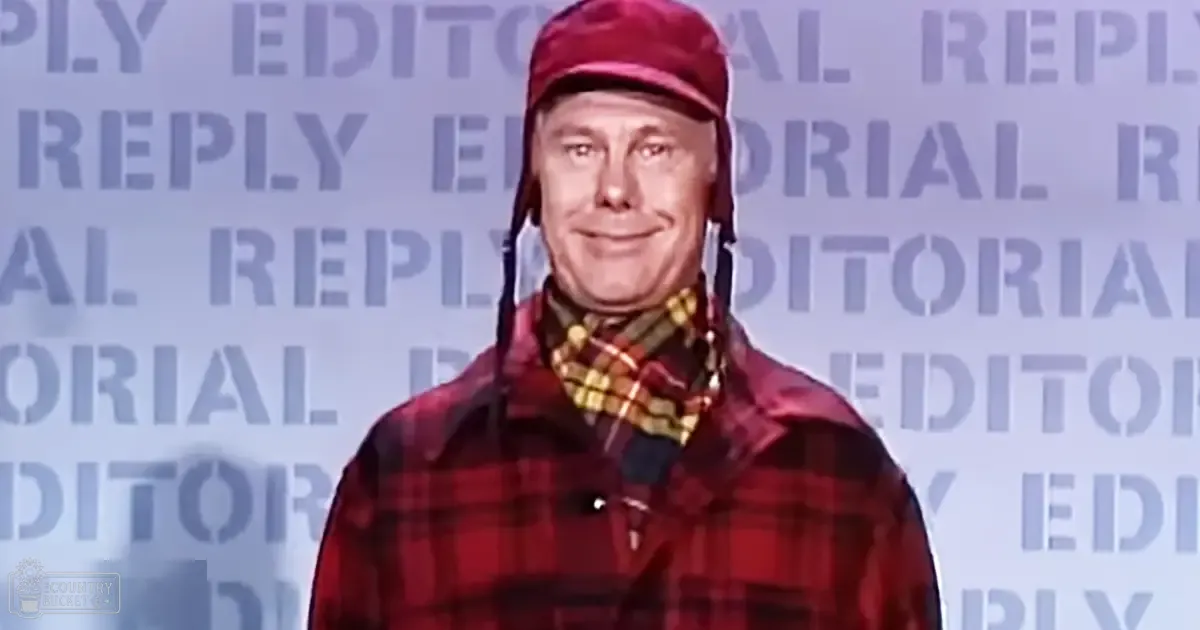 A middle-aged adult male wearing a red plaid jacket with long sleeves, casual style, and maroon cap indoors under professional stage lighting.