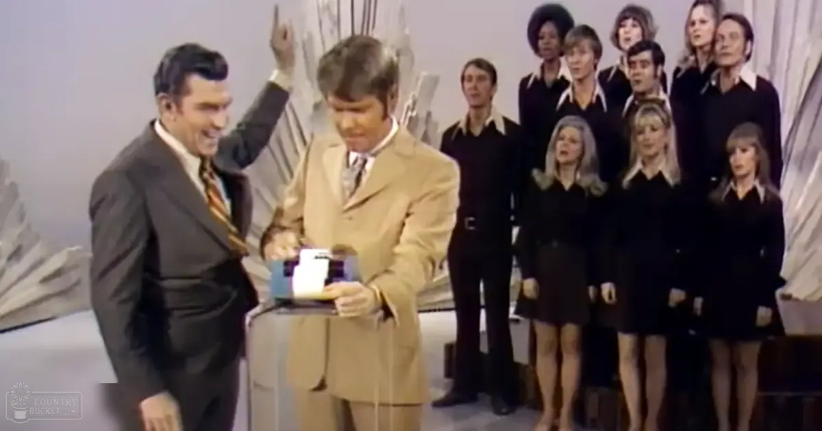 A man in a beige suit jacket stands alongside a young adult male in dark formal attire. They appear to be hosting or presenting something in a television studio setting with other adults and young adults dressed formally.
