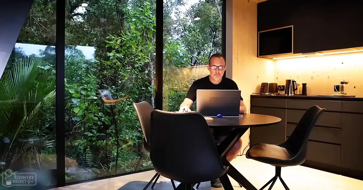 A modern kitchen interior with large windows showing lush greenery, suggesting a spring or summer setting during the daytime.