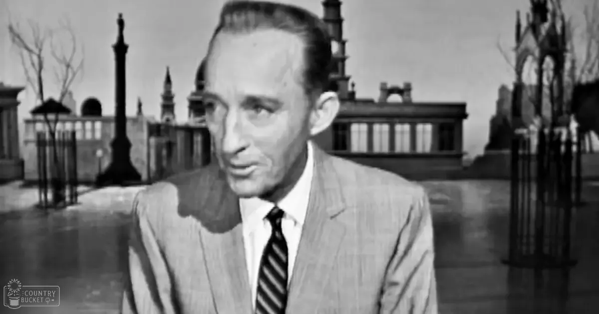An older adult male in a formal setting with a light-colored suit jacket, white shirt, and dark striped tie against an outdoor historical or classical building backdrop