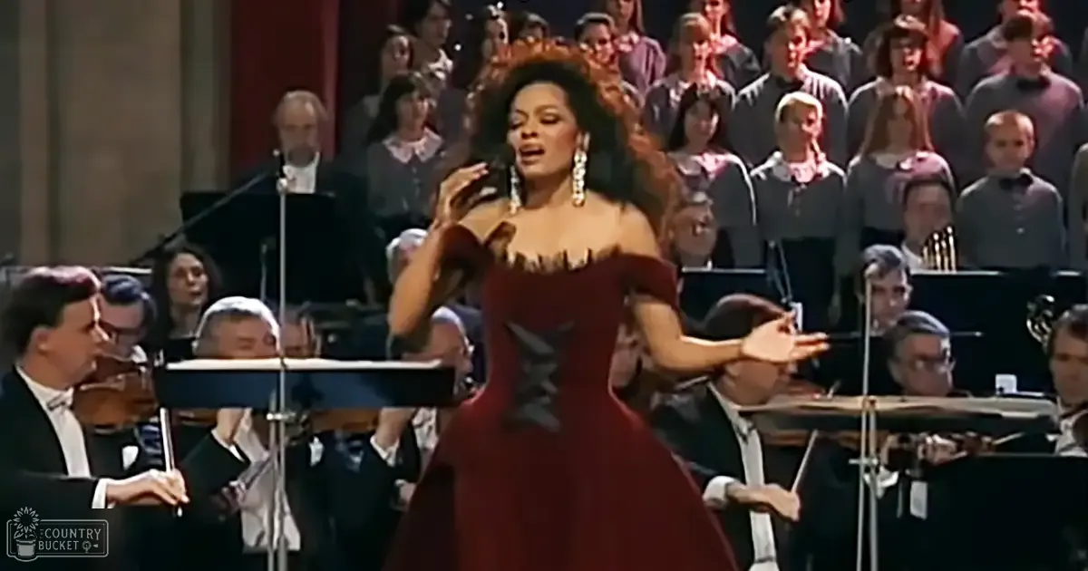 A woman in a deep red corset-style dress sings on stage, accompanied by an orchestra and choir at what appears to be a formal concert hall.