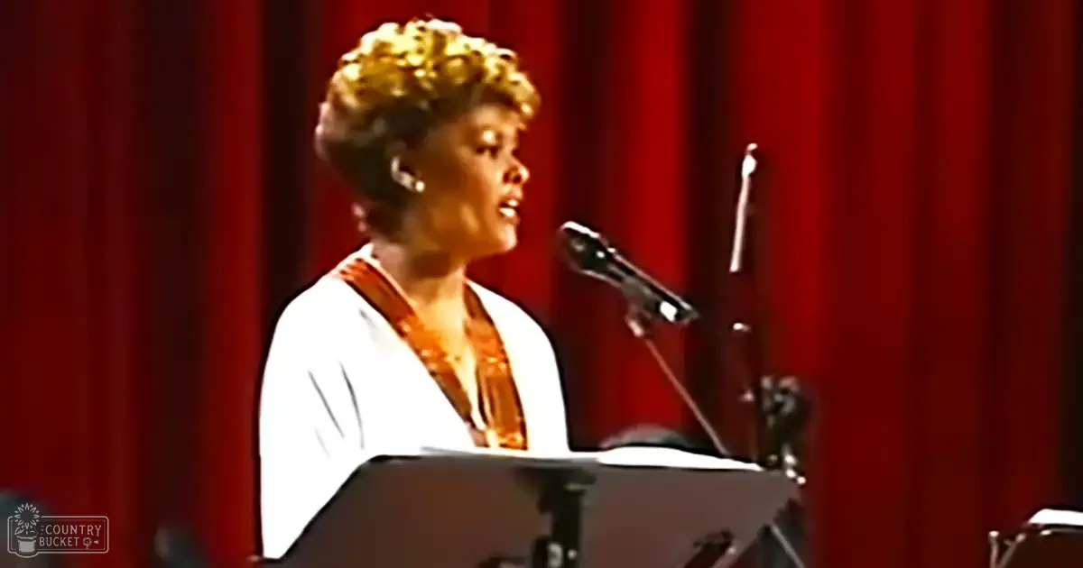 A female adult singer in a white v-neck top with orange-brown scarf, performing on stage with red curtains and microphone setup.